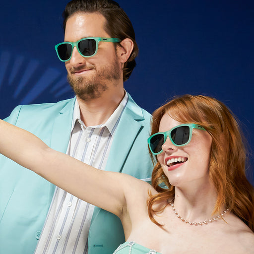 A woman and man in mint green formalwear and mint green wayfarer-shaped sunglasses with green gradient lenses smile.