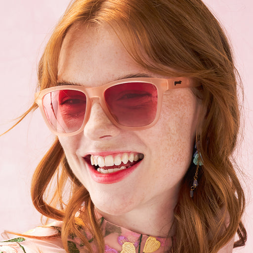 A smiling woman wears light pink sunglasses with rose-tinted gradient lenses and and matching floral dress.
