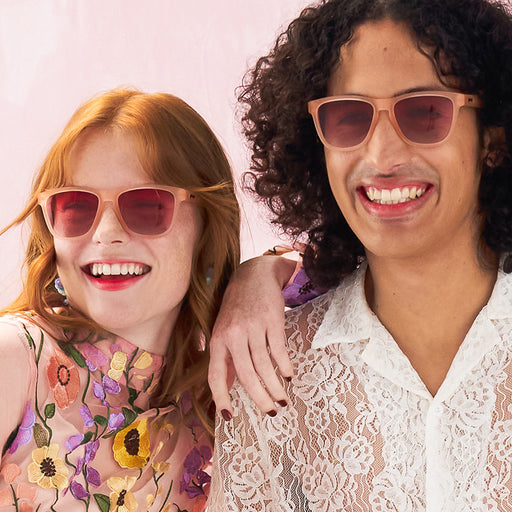 A man and woman smile in matching wayfarer-style blush pink sunglasses with gradient rose-tinted glasses.