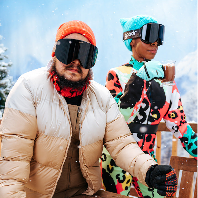 A man and woman in bright eccentric snow gear sip hot cocoa while wearing stylish black goodr snow goggles.