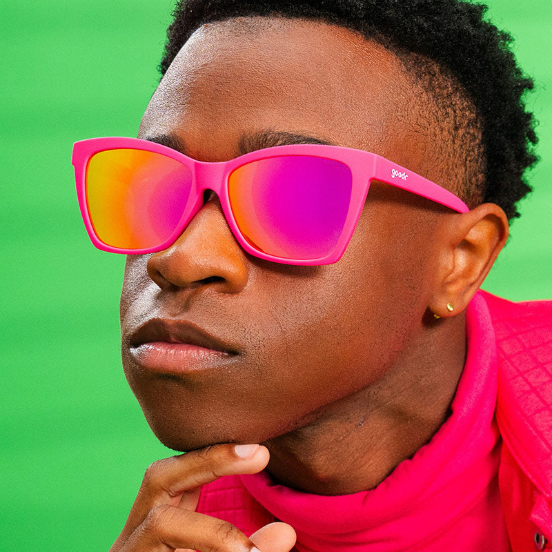 A man in a stylish hot pink outfit wears bright pink angled cat-eye sunglasses with reflective pink lenses.