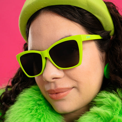 A woman in a trendy bright green outfit smiles in lime green angled cat-eye sunglasses with black gradient lenses.