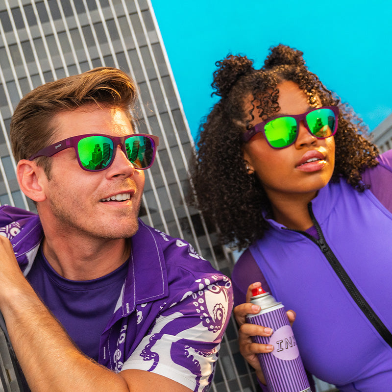 A man and woman in wide-fit purple sunglasses with green reflective lenses hold spray paint, eyeing miniature skyscrapers.