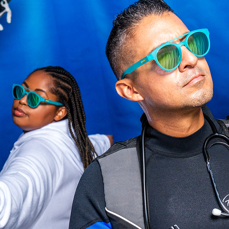 A man and woman in aqua blue double bridge sunglasses with aqua reflective frames stand before an ocean blue backdrop.