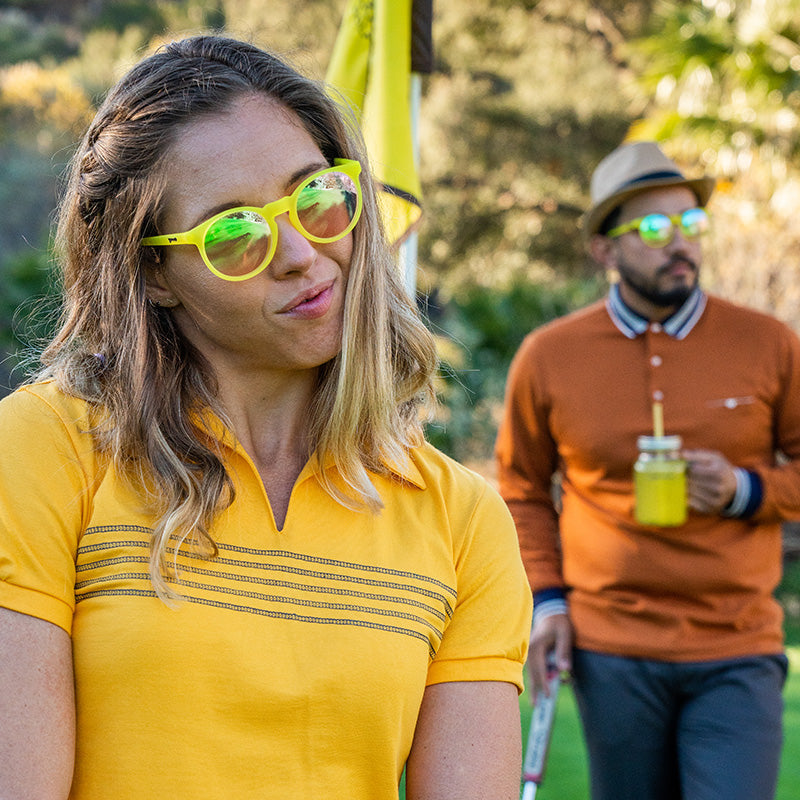A man sipping lemonade & a female golfer cheekily look out at a golf course in yellow sunglasses with round pink lenses.