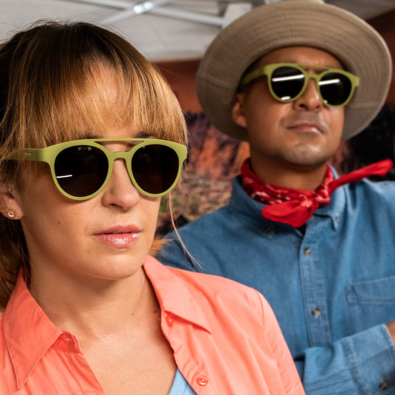 A woman and man wear iconic 90s archaeologist outfits & round olive green double-bridge sunglasses with brown lenses.