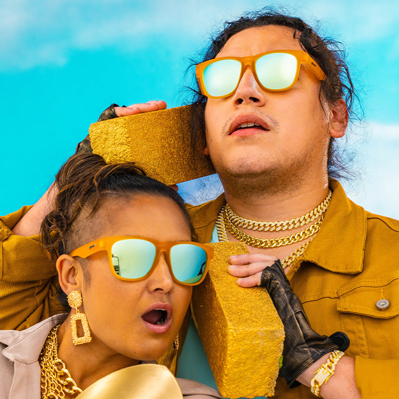 People in gold chains and orange square-shaped sunglasses with light blue mirrored frames hold gold bricks like phones.