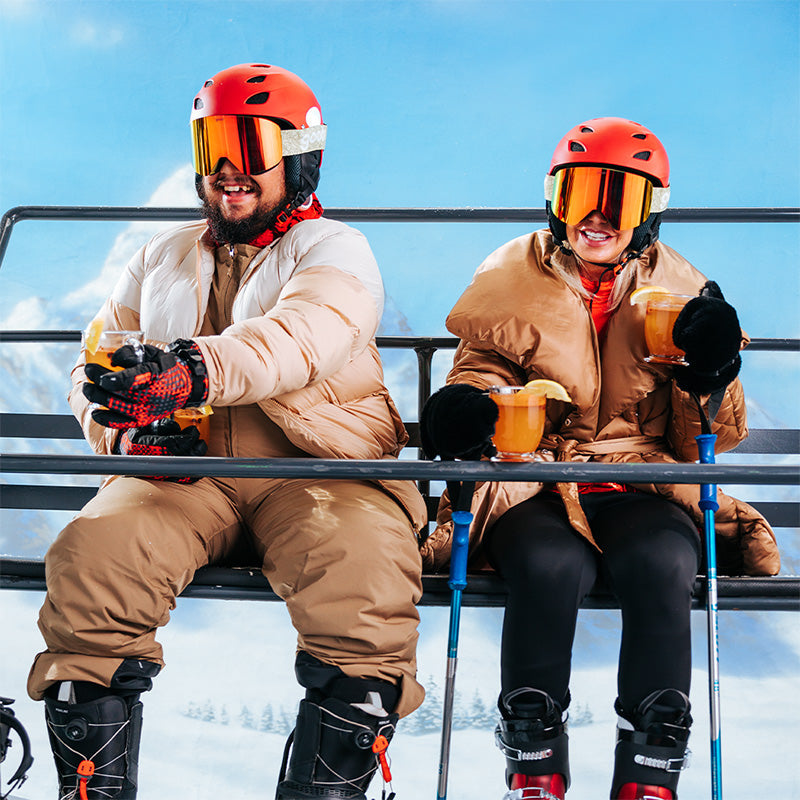 A male and female skier excitedly sip hot toddies on a chair lift wearing green snow goggles with a bright orange lens.