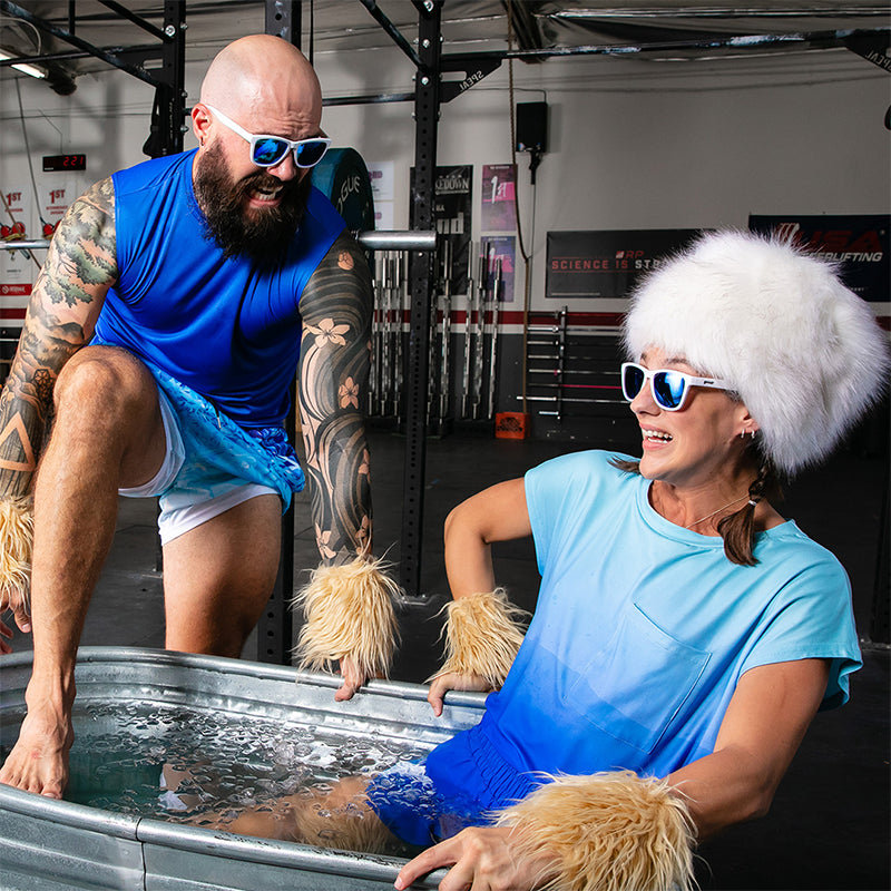 A tattooed man eases into an ice bath beside a woman in a white furry hat, both wearing white sunglasses with blue lenses.