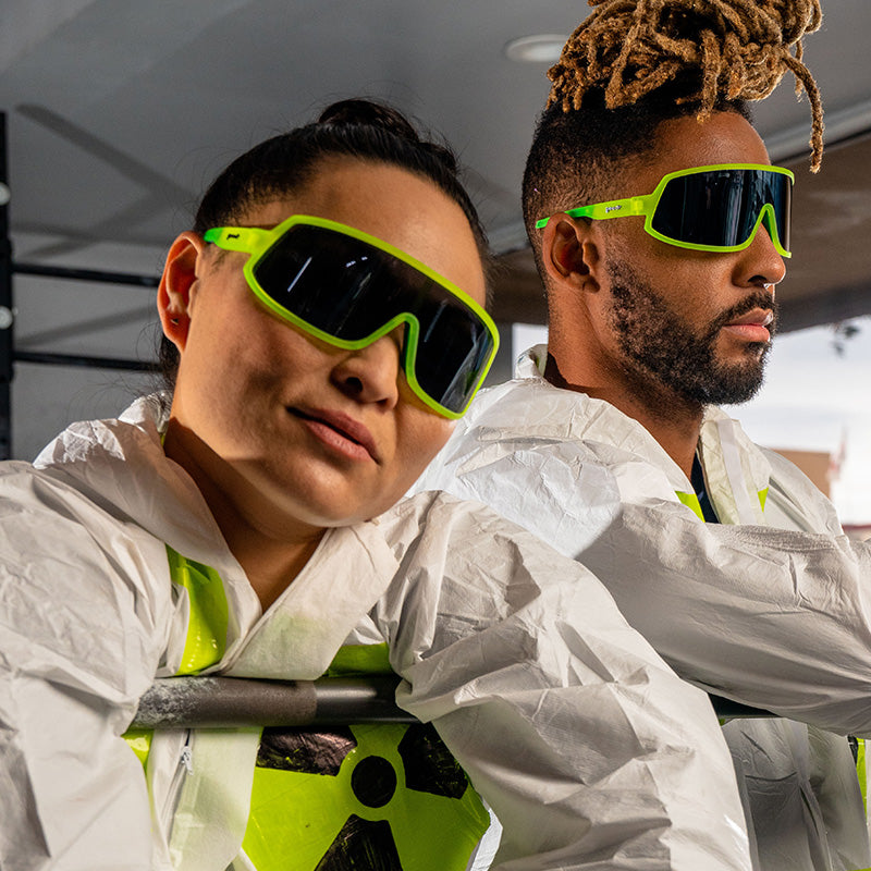 A man and woman in hazmat suits wear neon yellow sunglasses with a reflective dark gray lens.