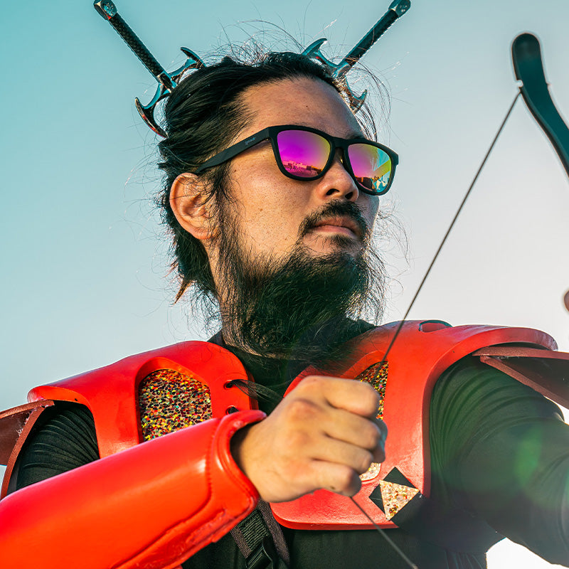 A man in a futuristic cosplay outfit and black square-shaped sunglasses with hot pink lenses looks ahead, holding a crossbow.
