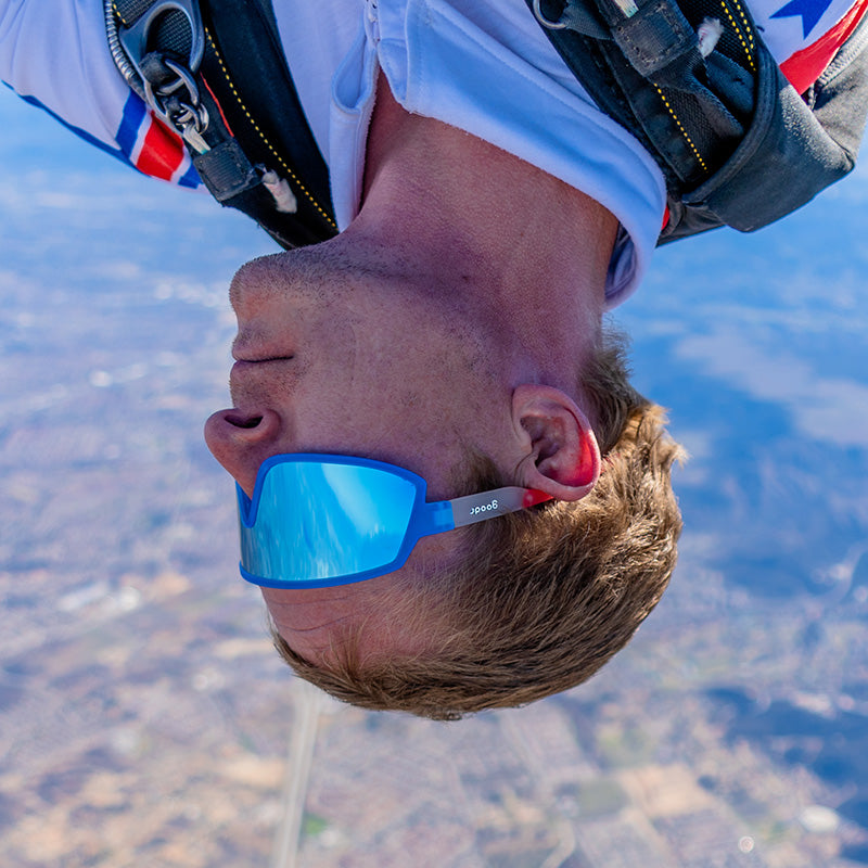 A skydiver wearing red, white, & blue wraparound sunglasses peeks out of an airplane, his sunglasses staying on his face.