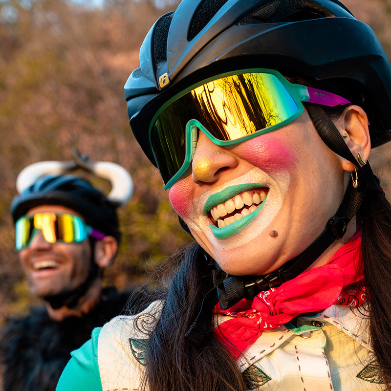 A female cyclist in teal & pink wraparound sunglasses & clown makeup stands before a male cyclist wearing bull horns.