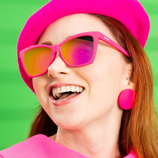 A smiling woman in a stylish hot pink outfit wears hot pink angled cat-eye sunglasses with pink reflective lenses.