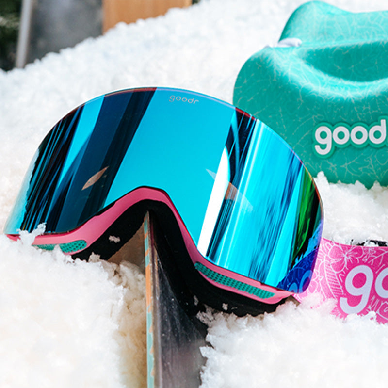 Three-quarter angle view of pink & blue snow goggles sitting on a snow bank with the teal protective case in the background.