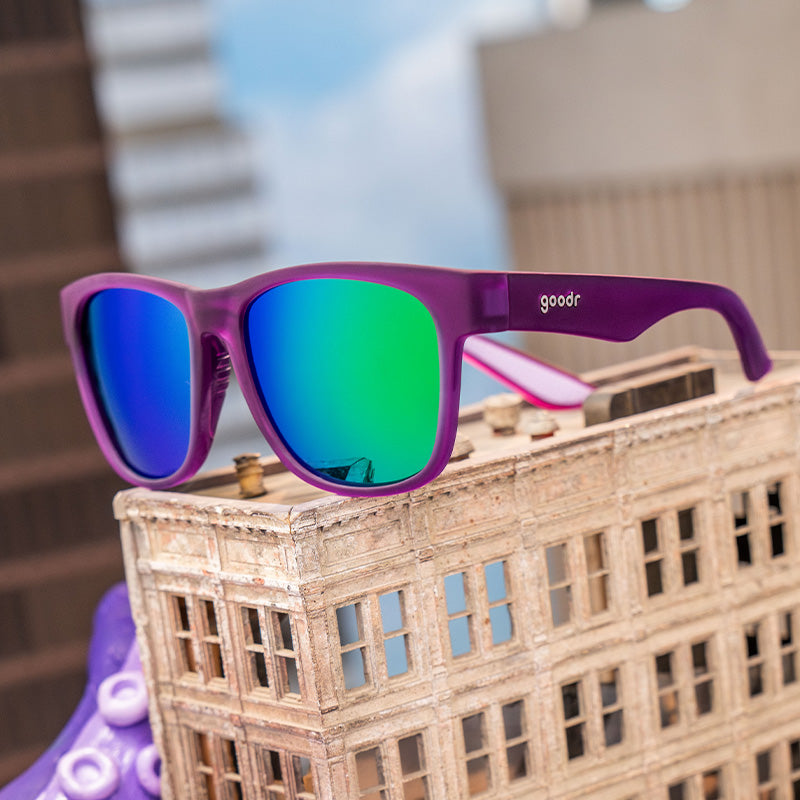 Three-quarter angle view of purple sunglasses with green reflective lenses on a mini skyscraper, a big tentacle in the back.