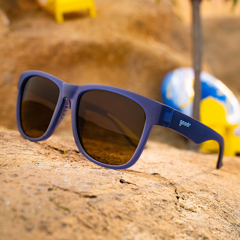 Three-quarter angle view of navy blue sunglasses with amber gradient lenses sitting atop a rock on a sandy shore.