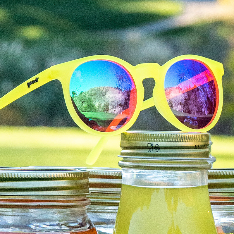 Three-quarter angle view of yellow golf sunglasses with pink round lenses sits atop jars of homemade lemonade.
