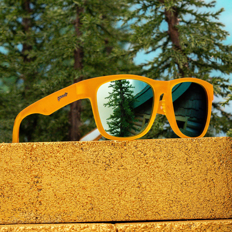 Three-quarter angle view of square-shaped orange sunglasses with light blue reflective lenses atop a gold-painted brick in a forest.