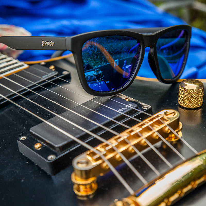 Three-quarter angle view of square-shaped black sunglasses with blue reflective lenses sitting atop an electric guitar.