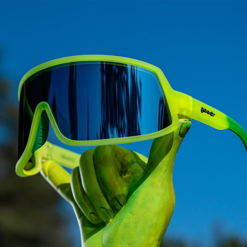 Three-quarter angle view of neon yellow wraparound sunglasses with a gray single lens balanced on a neon yellow painted hand.