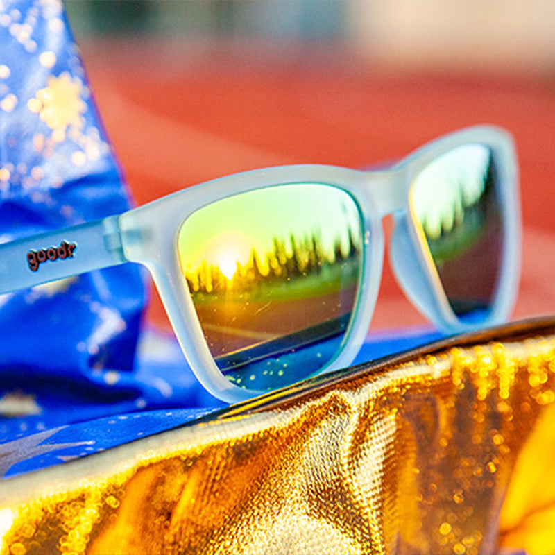 Three-quarter angle view of translucent light blue sunglasses with gold mirrored lenses sitting atop a wizard hat on a track.