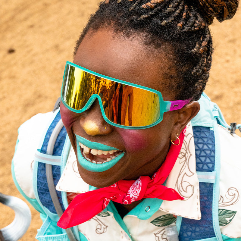 A female rodeo clown smiles wearing teal and pink wraparound sunglasses with a teal single lens.
