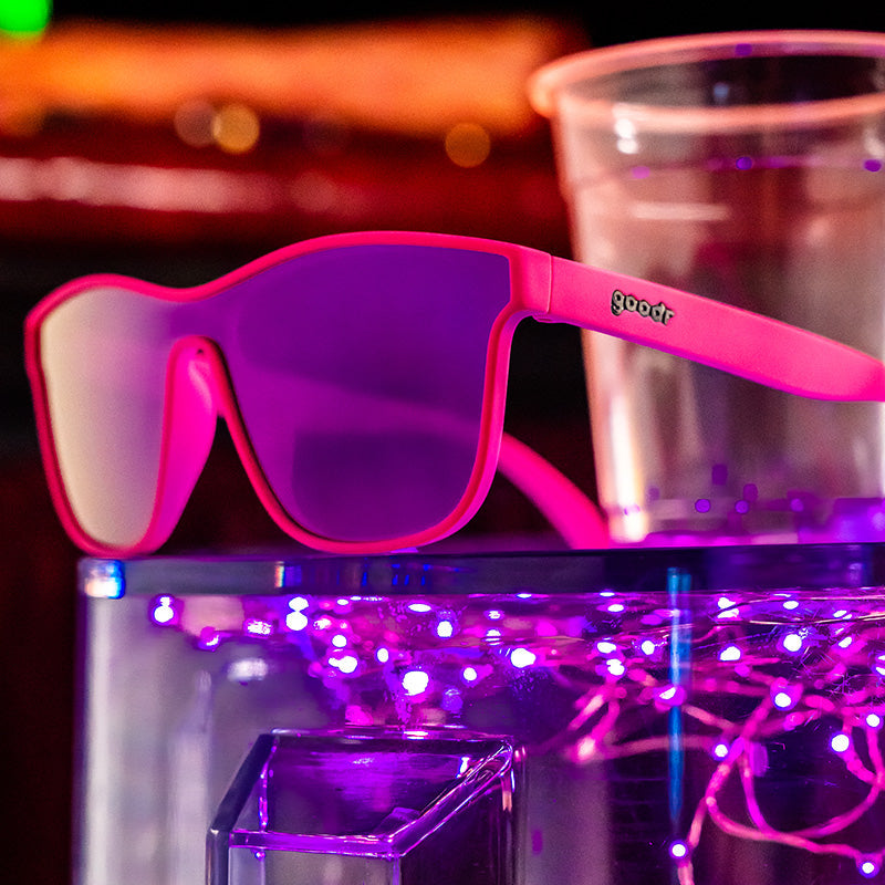 Three-quarter angle view of bright pink sunglasses with a futuristic reflective purple lens sitting atop a light-up bar.