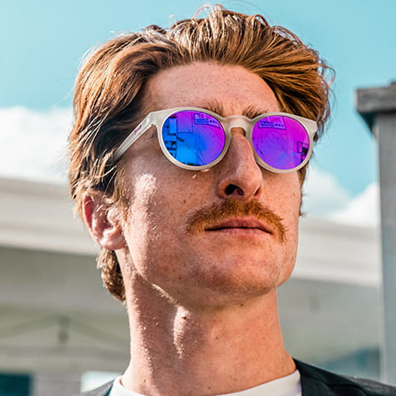 A three-quarter angle view of a man in circular clear sunglasses with round reflective purple lenses looks epically off.