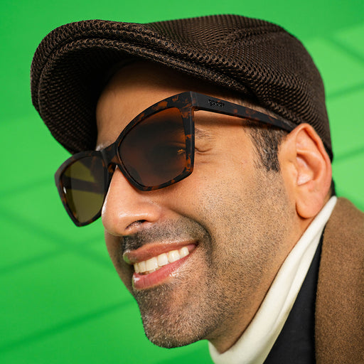 A man in a stylish brown outfit wears brown tortoiseshell angled cat-eye sunglasses, smiling off to the side.