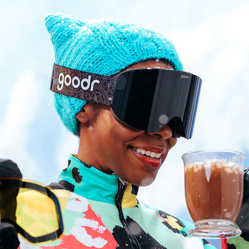 A smiling woman in bright snow gear and black snow goggles holds a magnetic swappable lowlight amber lens and hot cocoa.