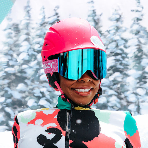 A woman in vibrant snow gear smiles to the side wearing pink & blue snow goggles with a pink strap and blue reflective lens.