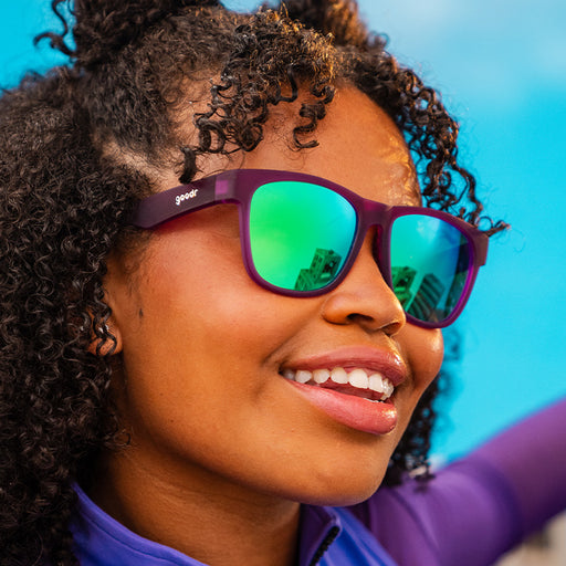 A woman in square-shaped purple sunglasses with green mirrored lenses looks out at a blue sky, smiling.