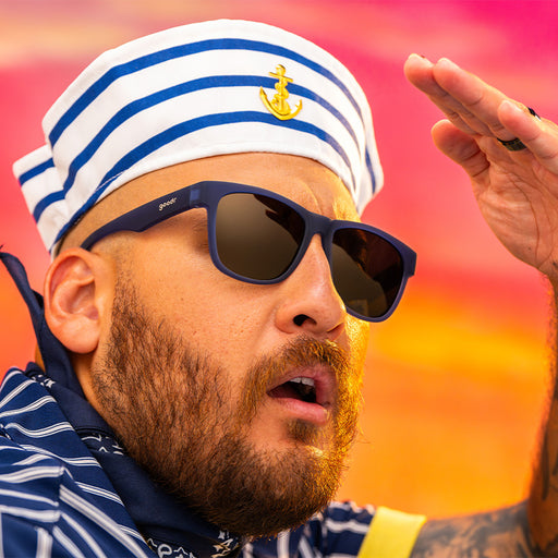 A man in a striped sailor cap and navy blue sunglasses with grey yellow gradient lenses excitedly looks into the horizon at sunset.