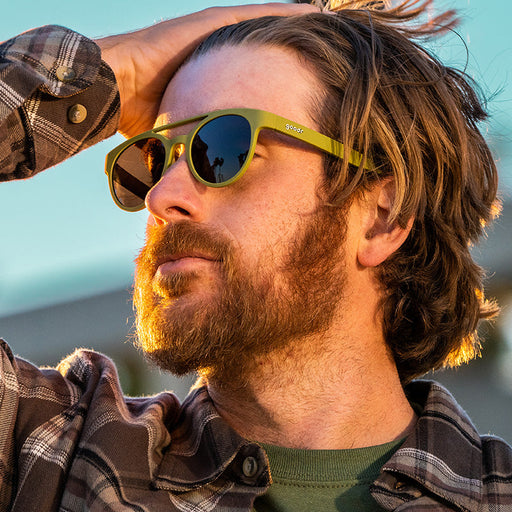 A man looking out at a sunset wears olive green double bridge round sunglasses with brown non-reflective lenses.