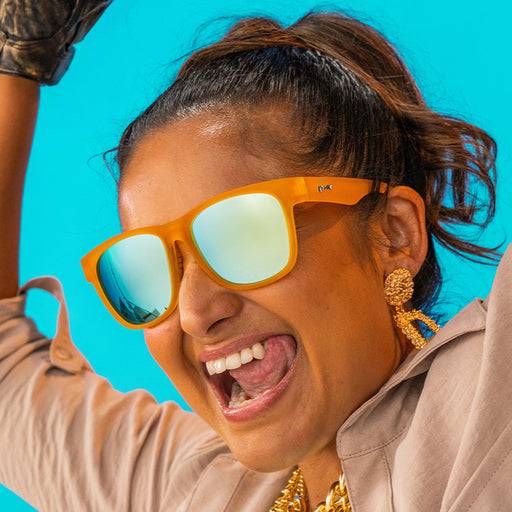 A woman in orange sunglasses with light blue mirrored lenses and chunky gold jewelry licks her chops on a sunny day.