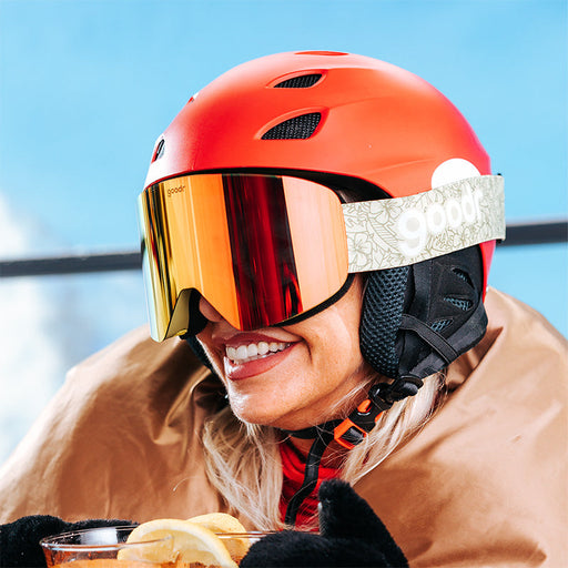 A smiling woman sips a hot toddy on a chair lift in vibrant snow goggles with a orange reflective lens and olive green strap.