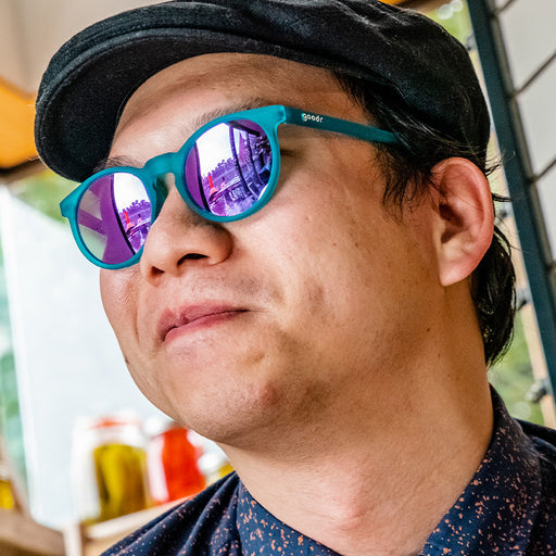 A man wearing circle-shaped teal sunglasses with round purple lenses looks off to the side confidently.