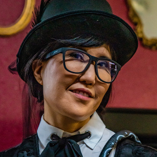 A woman in black square-shaped sunglasses with clear blue-light-blocking lenses smugly smiles in a top hat.