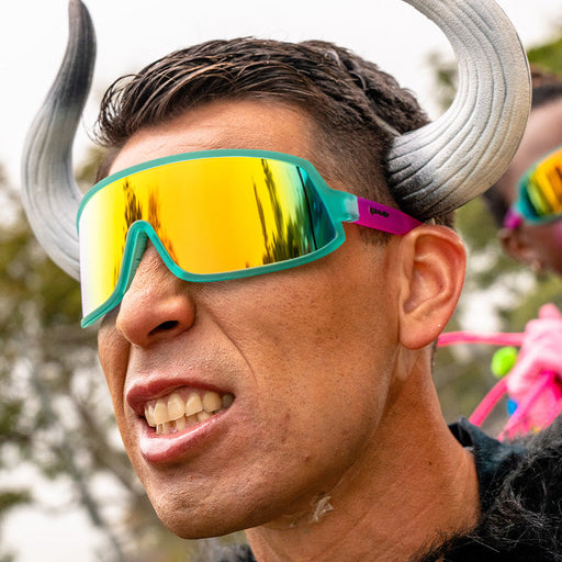 A man wearing bull horns and stylish teal and pink wraparound sunglasses fiercely bears his teeth.