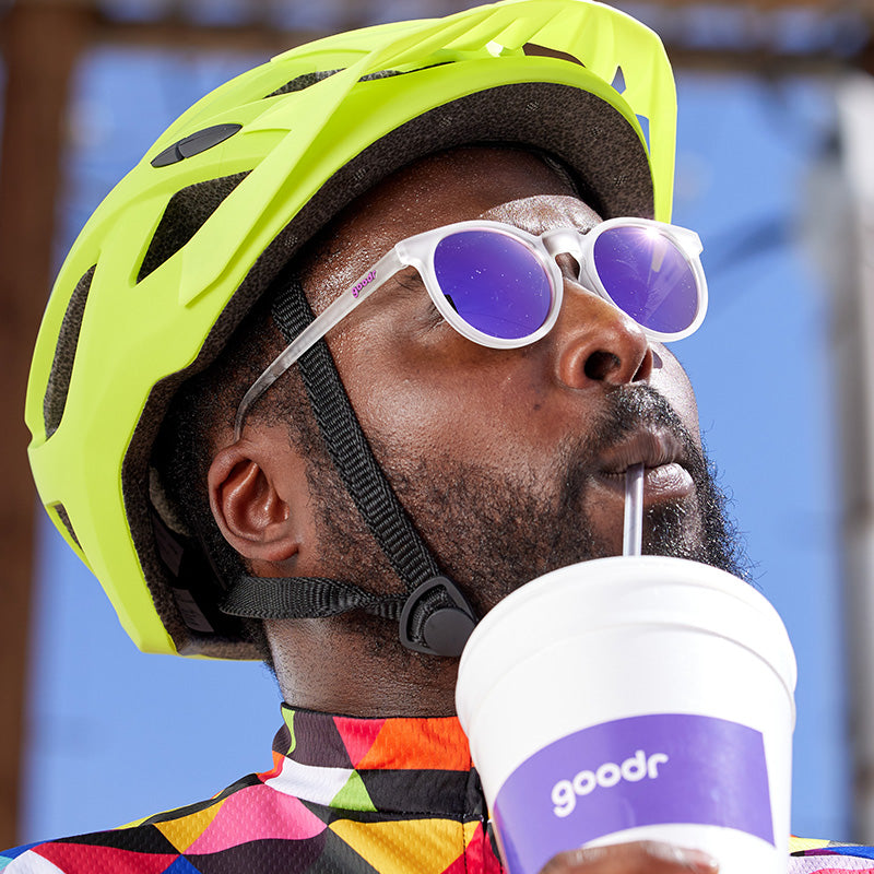 A man in a neon yellow helmet and clear sunglasses with round purple lenses cheerfully sips a giant gas station drink.