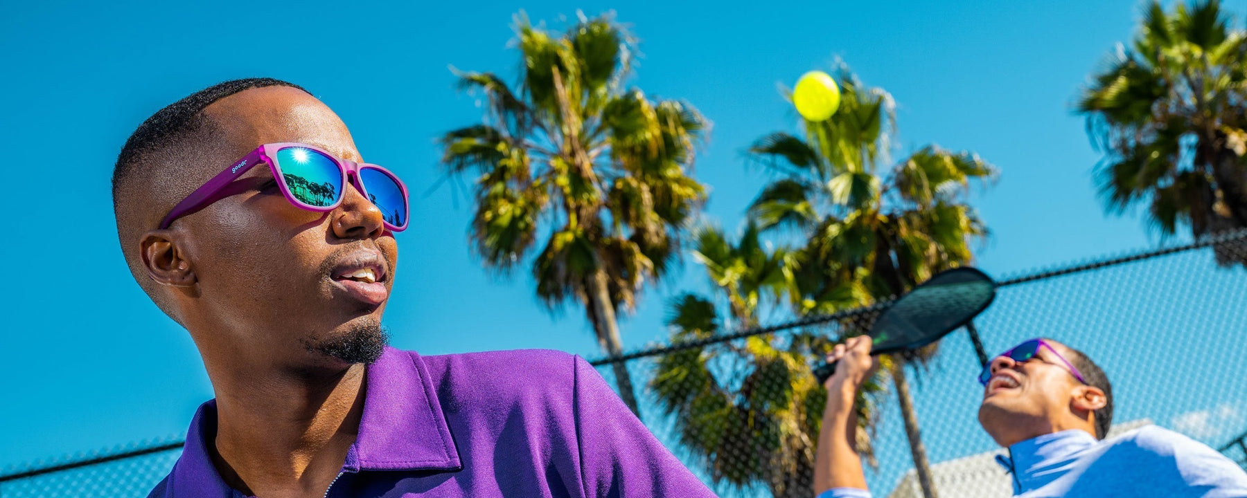 Person wearing purple goodr sunglasses playing tennis outside.