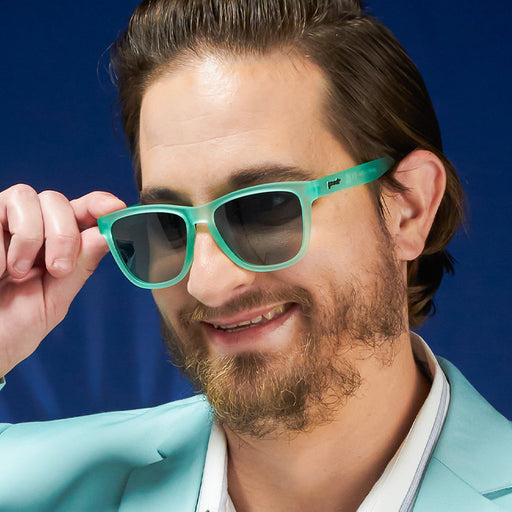 A smiling man wears mint green wayfarer-style sunglasses with green gradient lenses and a matching mint suit.