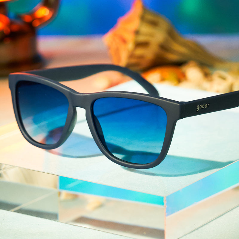 Three-quarter angle view of navy blue sunglasses with gradient blue lenses sitting atop an acrylic display.