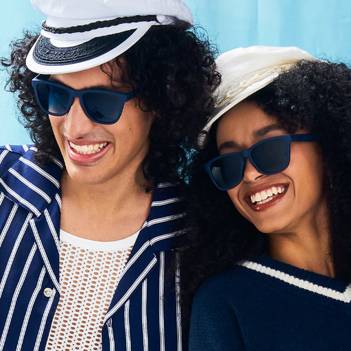 A smiling man and woman wear navy blue sunglasses with blue lenses and coordinating stylish nautical outfits.