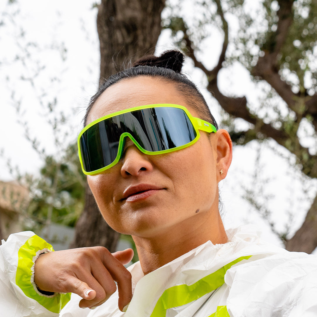 A woman wearing a hazmat suit and neon yellow sunglasses with a reflective gray lens looks fiercely ahead.