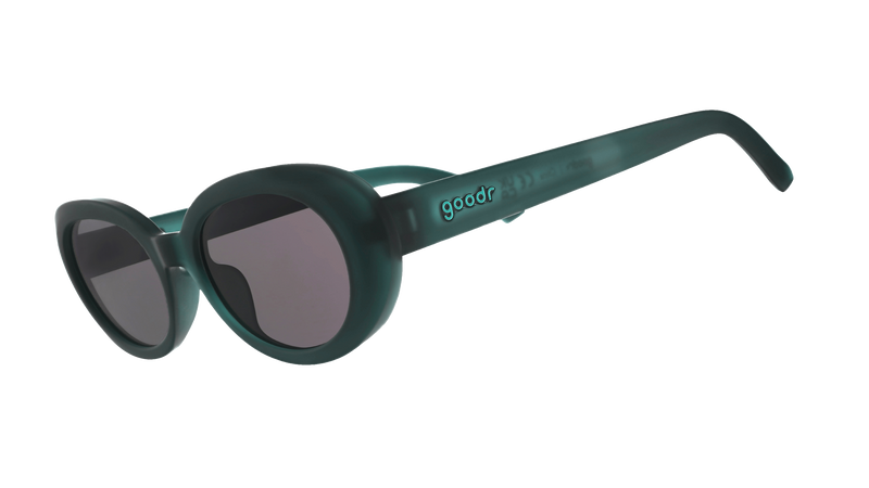Beat G oval sunglasses with a matte dark green frame and dark lenses, resting on a colorful patchwork knit cardigan with a navy blue ribbed beanie partially visible at the top.