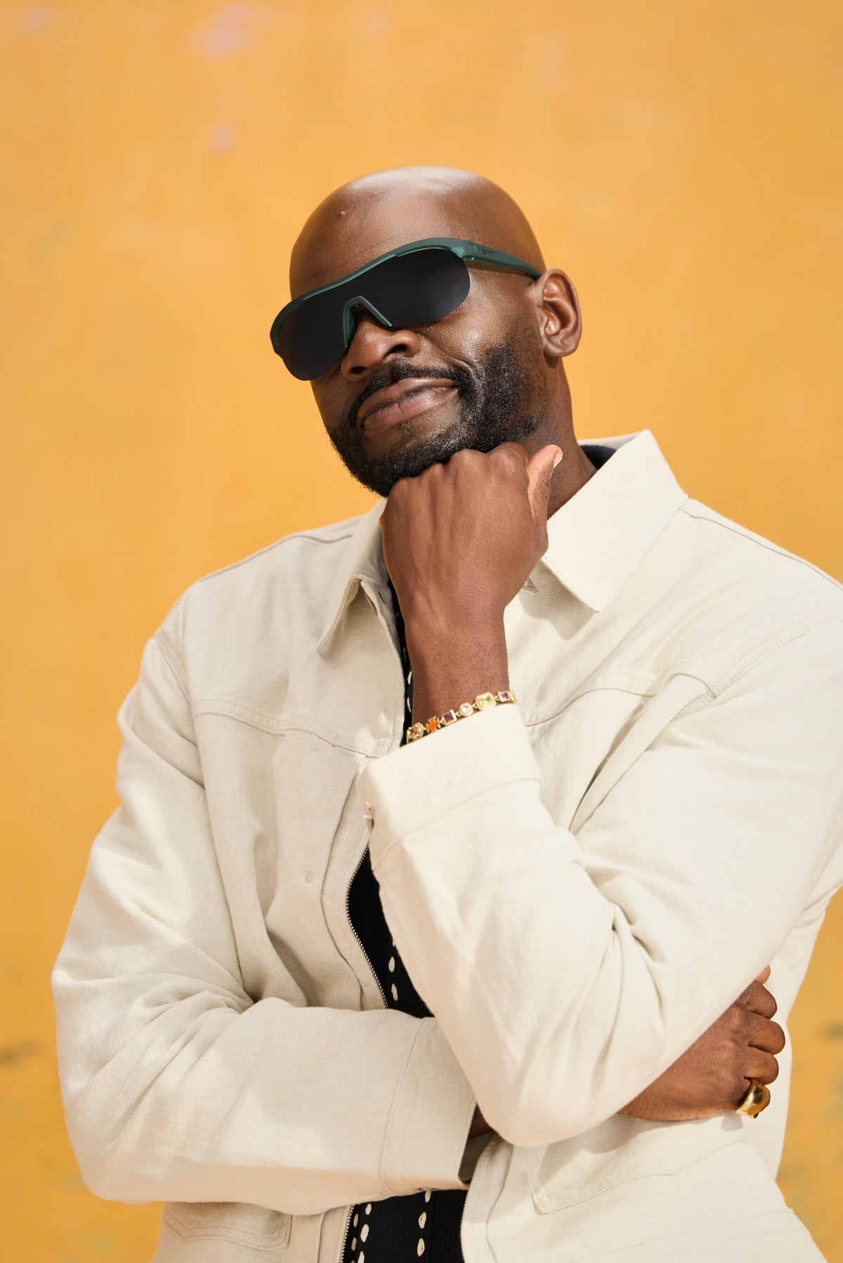 A man wearing goodr sunglasses with a green frame, a cream-colored jacket, and a black shirt, posing against a textured yellow background.