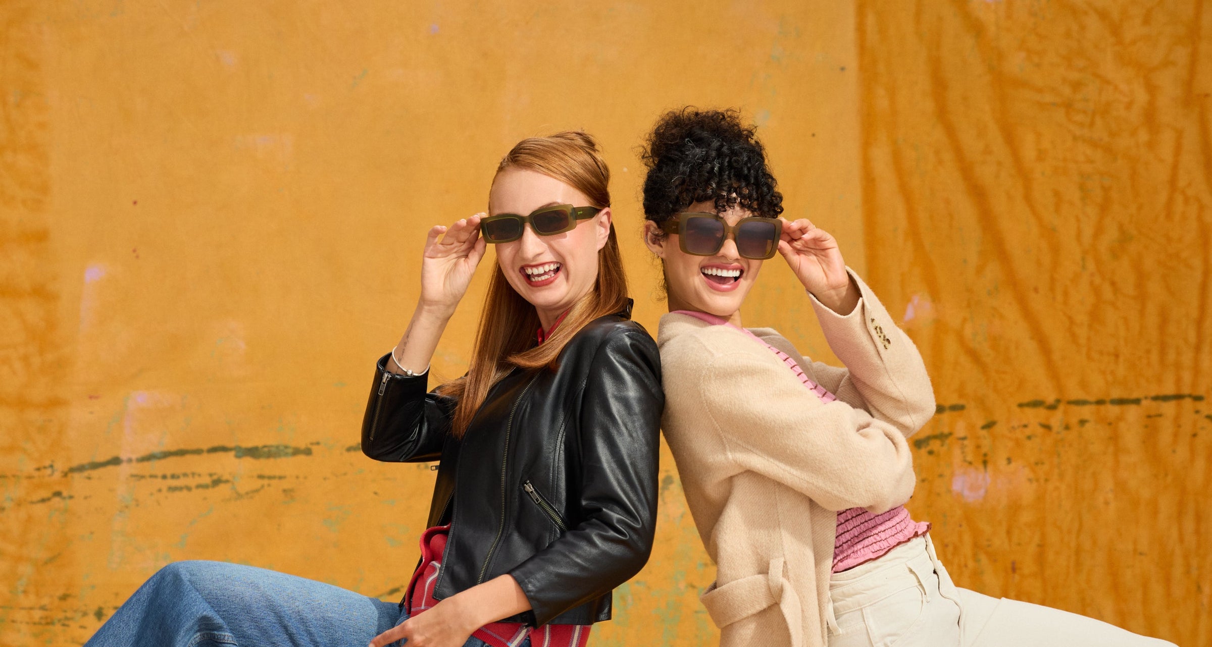 Two women posing back-to-back in stylish goodr sunglasses from the Fall Winter 2026 Collection, a black leather jacket, blue jeans, and a beige trench coat against a textured orange wall.