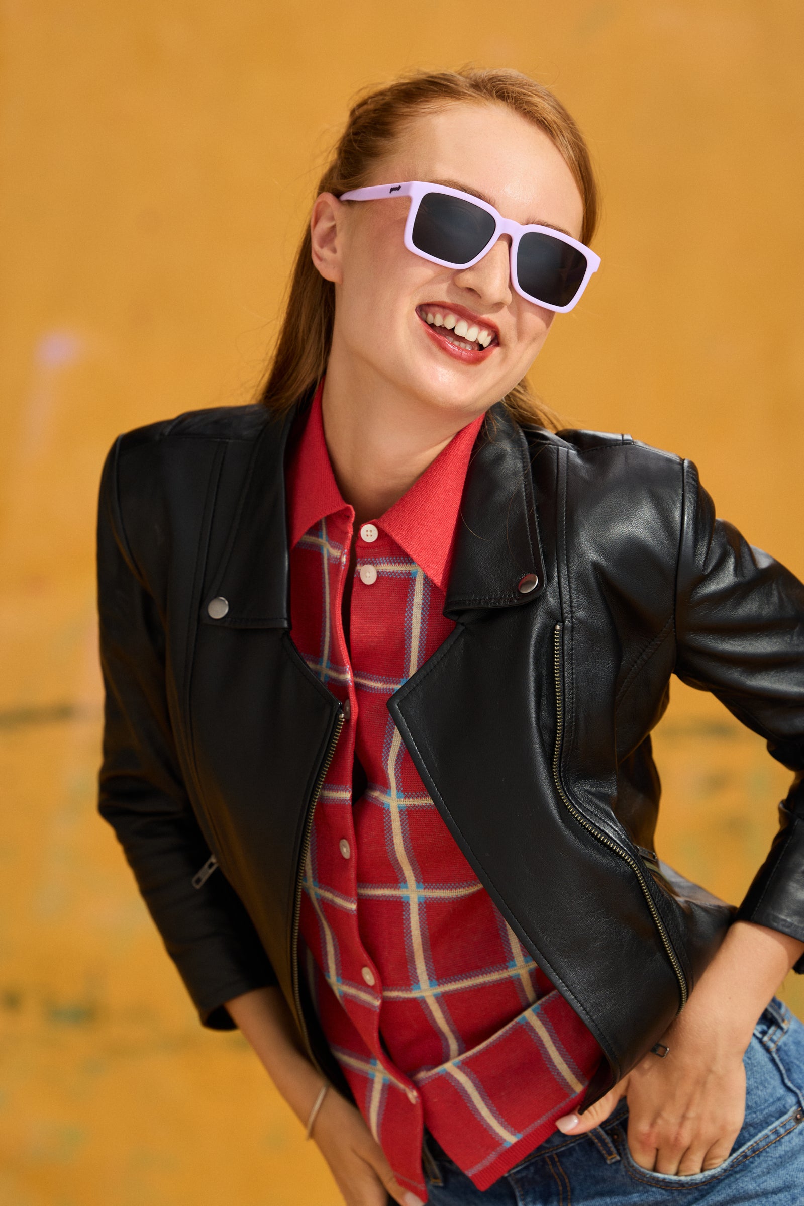 A woman wearing a black leather jacket over a red plaid button-up shirt, paired with blue jeans and light purple Slate G goodr square sunglasses, posing against a textured yellow background.